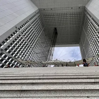 Broad stone steps lead up beneath a monumental open-sided building with gridded windows and a suspended canopy.