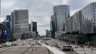 Modern glass-and-steel high-rise buildings surround a broad pedestrian plaza beneath a cloudy sky.