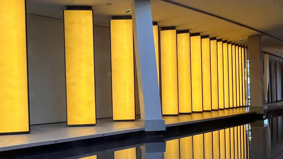 A row of tall glowing yellow rectangular light panels lines an indoor corridor beside a dark reflective pool.