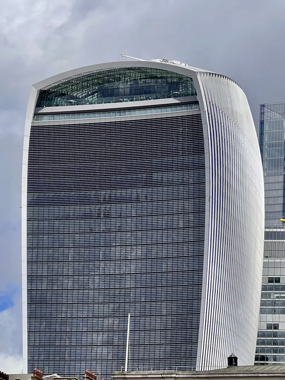 A tall modern office tower with a curved white frame and a dark louvered glass facade rises against a cloudy sky.