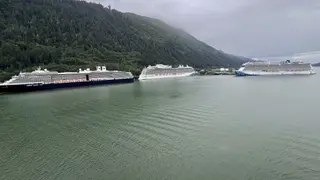 Three large cruise ships are docked along a calm body of water beneath a densely forested mountainside.