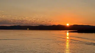 The setting sun casts a bright orange reflection across calm water beneath silhouetted hills.