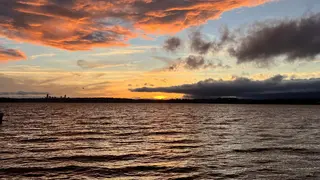 Orange and gray clouds glow above rippling water as the sun sets behind a dark shoreline.