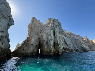 Jagged pale rock cliffs rise from clear turquoise ocean water beneath a cloudless blue sky.