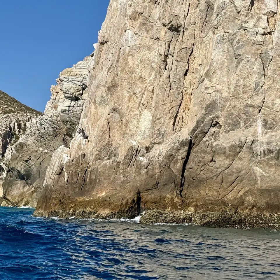 A towering weathered rocky cliff rises directly from deep blue ocean water beneath a clear sky.