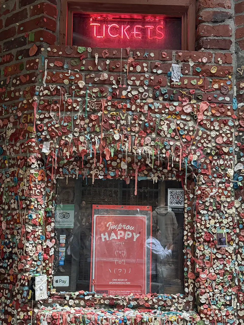 A red neon "TICKETS" sign glows above a window and brick wall densely covered with multicolored chewing gum.