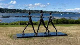 Three black statues of people walking on a black tile platform in front of a body of water.
