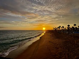 People walk along a sandy beach under a cloudy sky at sunset.