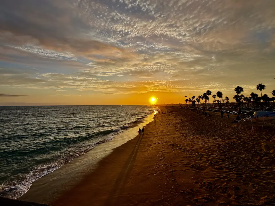 People walk along a sandy beach under a cloudy sky at sunset.