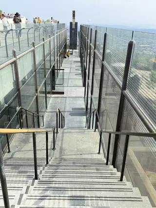 A staircase with glass walls and metal railings leads down to a rooftop terrace with people standing on it.