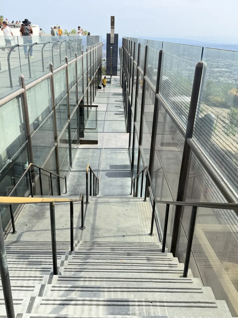 A staircase with glass walls and metal railings leads down to a rooftop terrace with people standing on it.