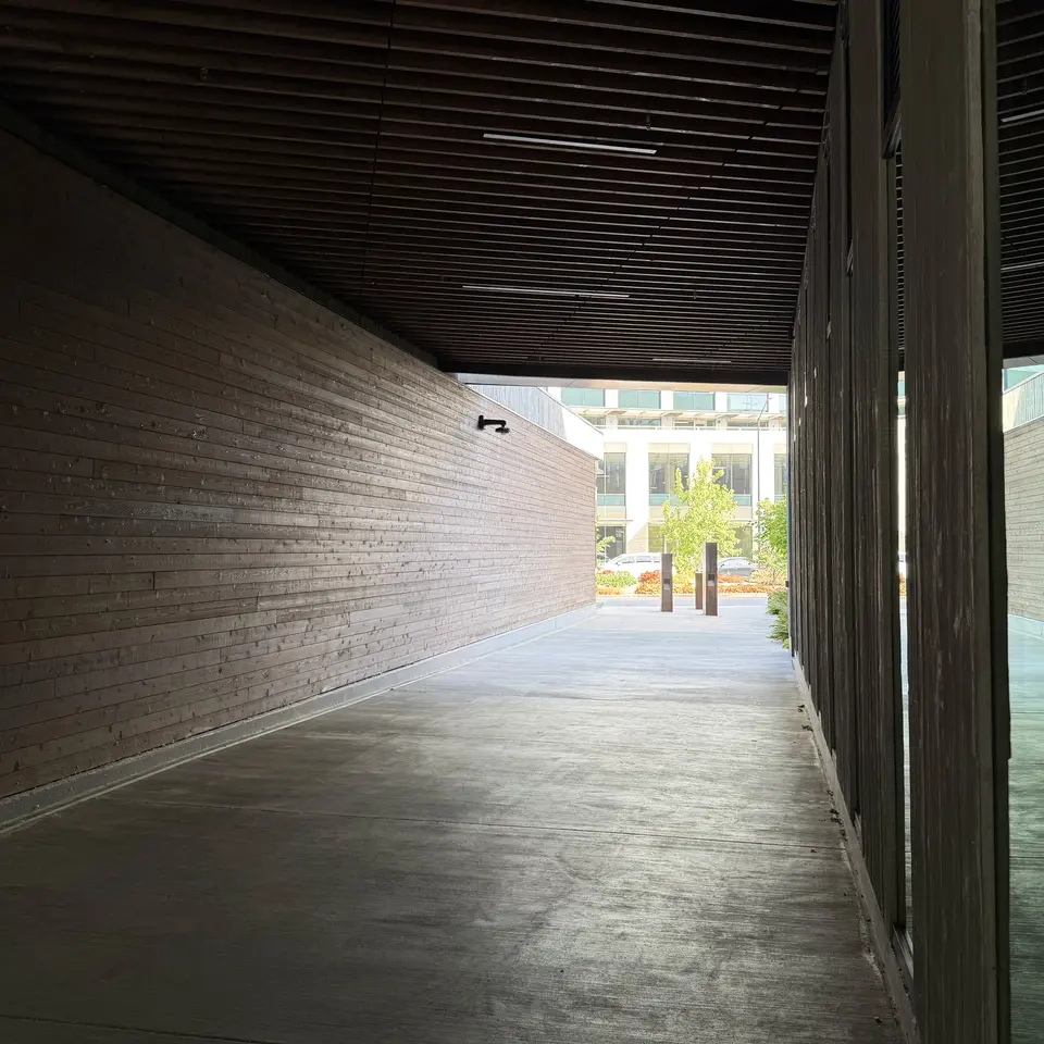 A covered concrete walkway with a brick wall, slatted ceiling, and glass panels opens onto a sunlit courtyard.