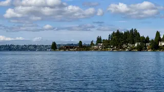 A calm expanse of rippling water stretches toward a tree-lined residential shoreline beneath scattered clouds.