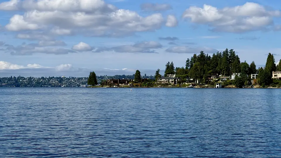 A calm expanse of rippling water stretches toward a tree-lined residential shoreline beneath scattered clouds.