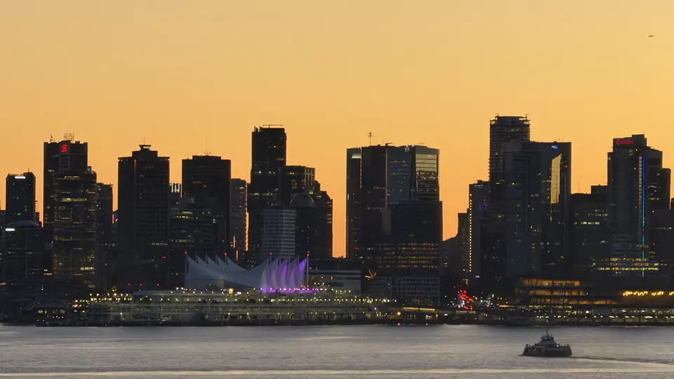 Vancouver’s skyline and the sail-shaped Canada Place stand silhouetted against an orange sunset above the water, with a small boat in the foreground.