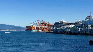 Canada Place and a cruise ship stand beside container cranes and a cargo vessel on Burrard Inlet beneath a clear blue sky.