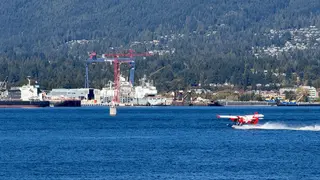 A red seaplane skims across blue water in front of cranes, ships, and a forested hillside.