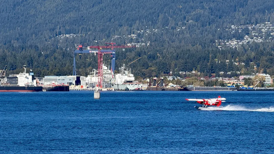 A red seaplane skims across blue water in front of cranes, ships, and a forested hillside.