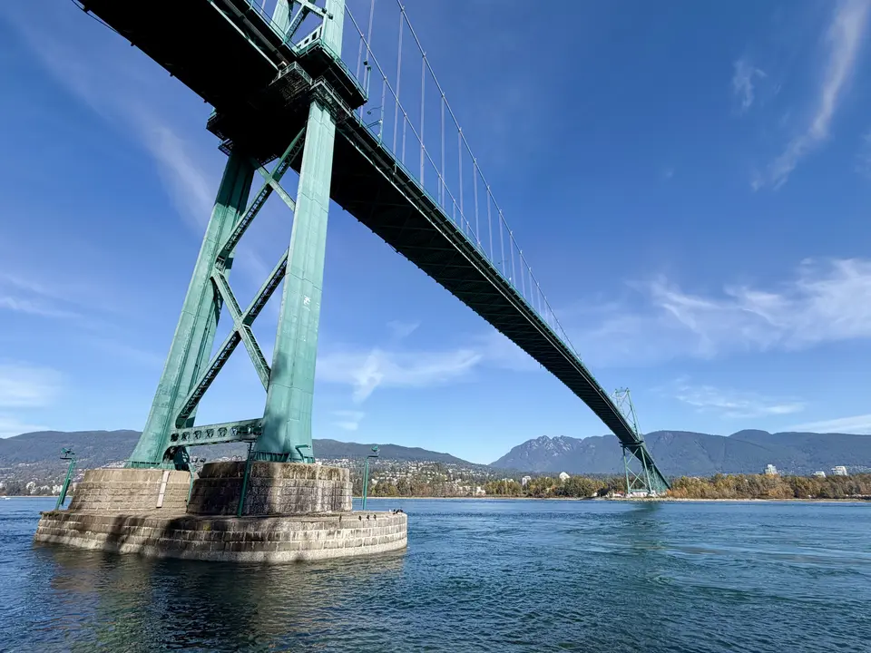 The Lions Gate Bridge spans blue water beneath a clear sky, with forested mountains in the distance.