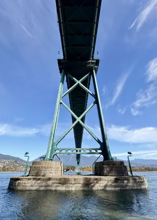 The Lions Gate Bridge rises overhead on green steel supports above the water, with mountains in the distance beneath a blue sky.