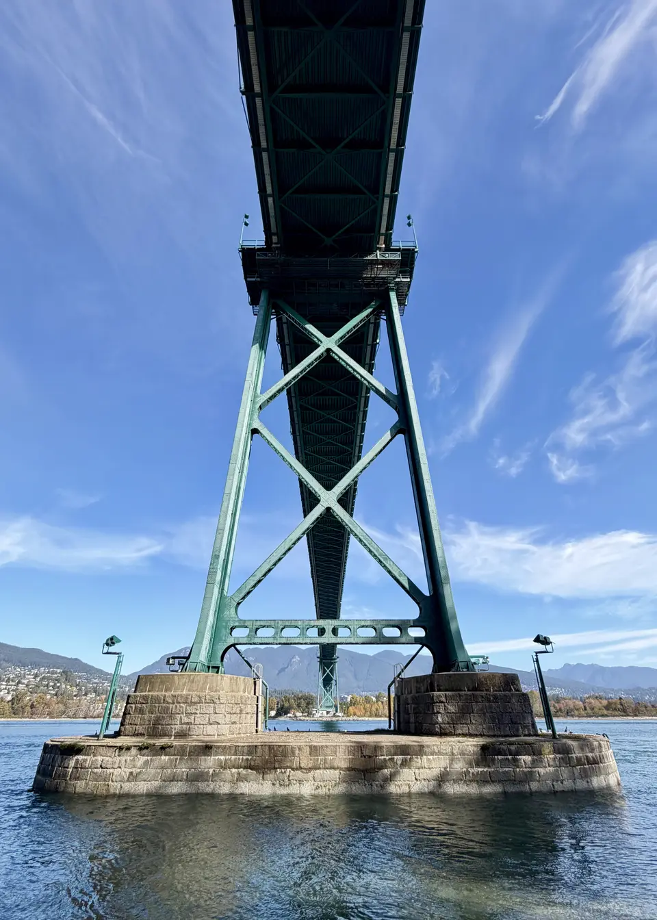 The Lions Gate Bridge rises overhead on green steel supports above the water, with mountains in the distance beneath a blue sky.
