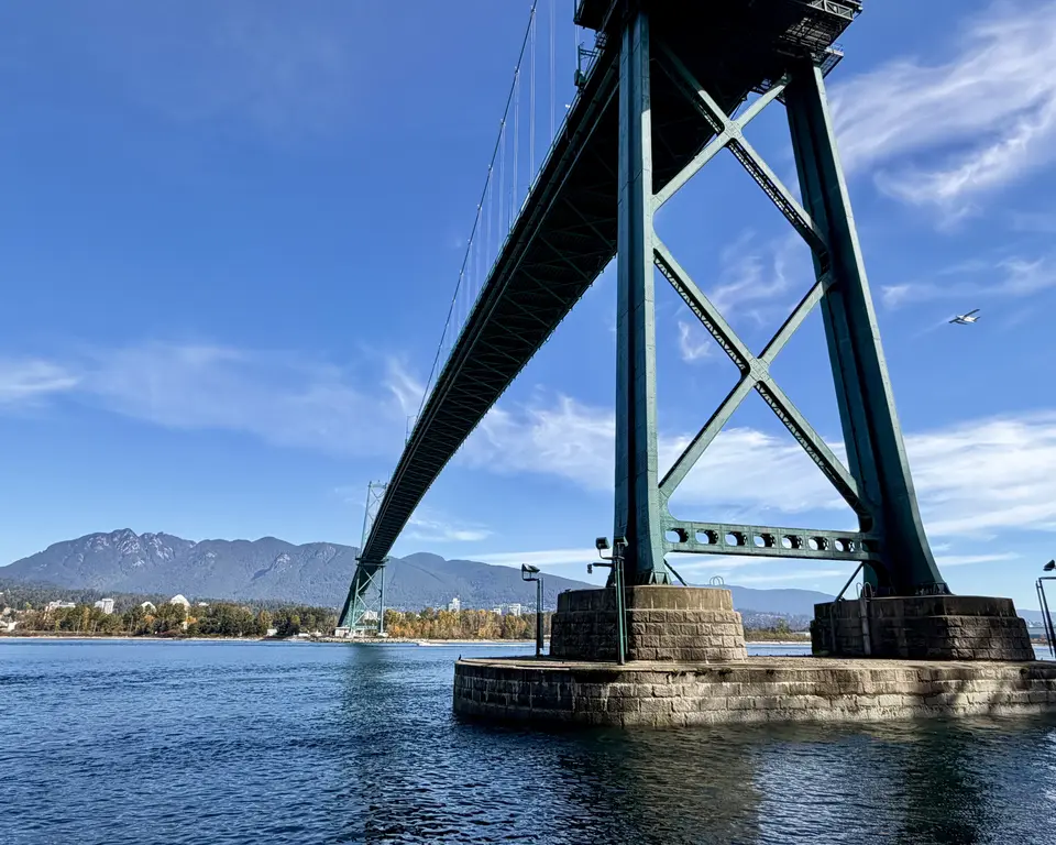 The Lions Gate Bridge spans blue water beneath a bright sky, with mountains and a shoreline in the distance.