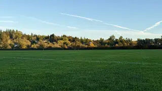 A broad green grass field stretches toward autumn-colored trees and scattered houses beneath a clear blue sky.