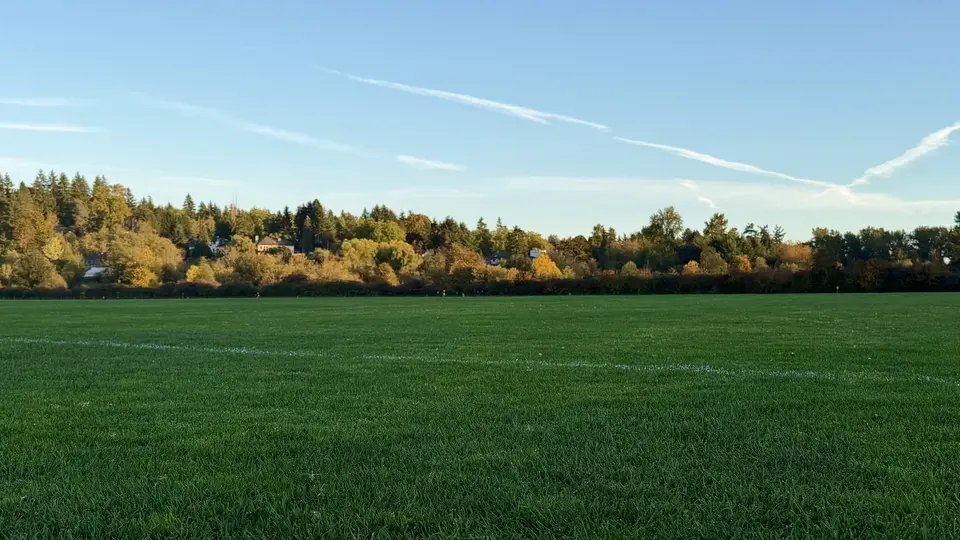 A broad green grass field stretches toward autumn-colored trees and scattered houses beneath a clear blue sky.