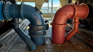 Large red and blue metal pipes with bolted joints stand beneath a covered industrial structure beside the water.