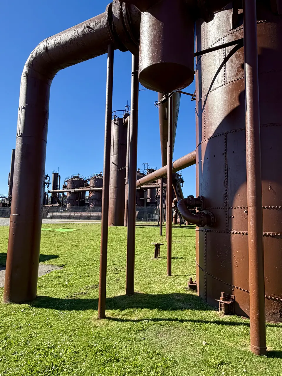 Rust-colored industrial pipes and riveted tanks stand above a grassy lawn beneath a clear blue sky.