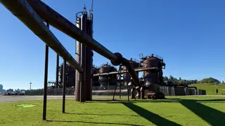 Rusting industrial pipes and towers at Gas Works Park rise above a sunlit lawn beneath a clear blue sky.