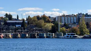 Colorful floating homes and a moored boat line the waterfront below hillside houses and apartment buildings under a blue sky.