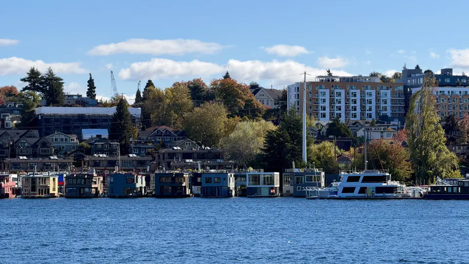 Colorful floating homes and a moored boat line the waterfront below hillside houses and apartment buildings under a blue sky.